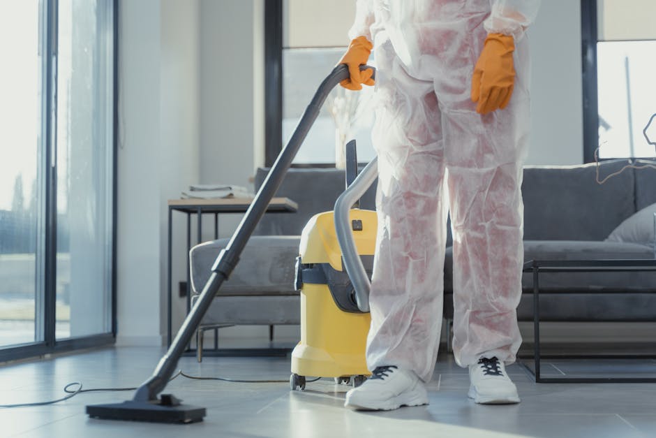 A cleaning professional from Cleaning Home is performing deep cleaning in a modern living room, using a yellow and grey vacuum cleaner. The individual is wearing protective white coveralls and orange 