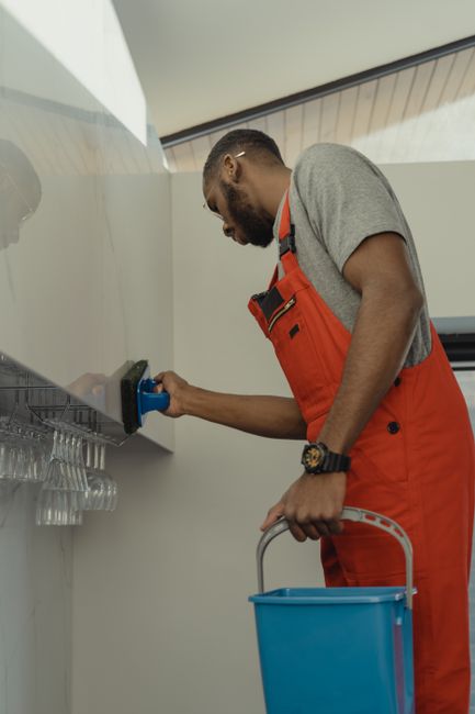 A cleaning professional from Cleaning Home is performing deep cleaning in a modern living room, using a yellow and grey vacuum cleaner. The individual is wearing protective white coveralls and orange 