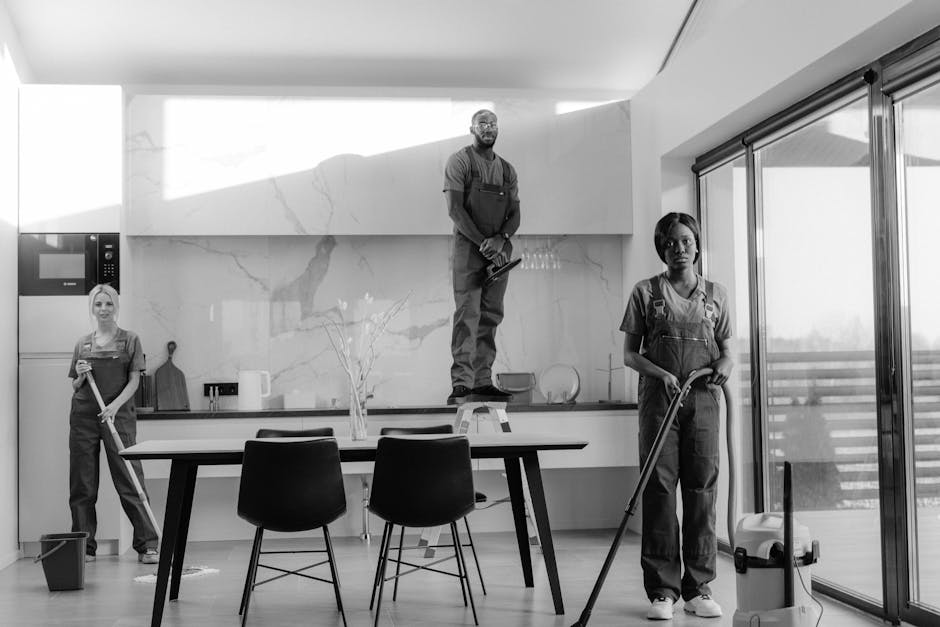 Three professional cleaners dressed in red uniforms are walking up the steps of a modern, two-storey house with a combination of white and wood-paneled exterior walls. One is holding a vacuum cleaner,